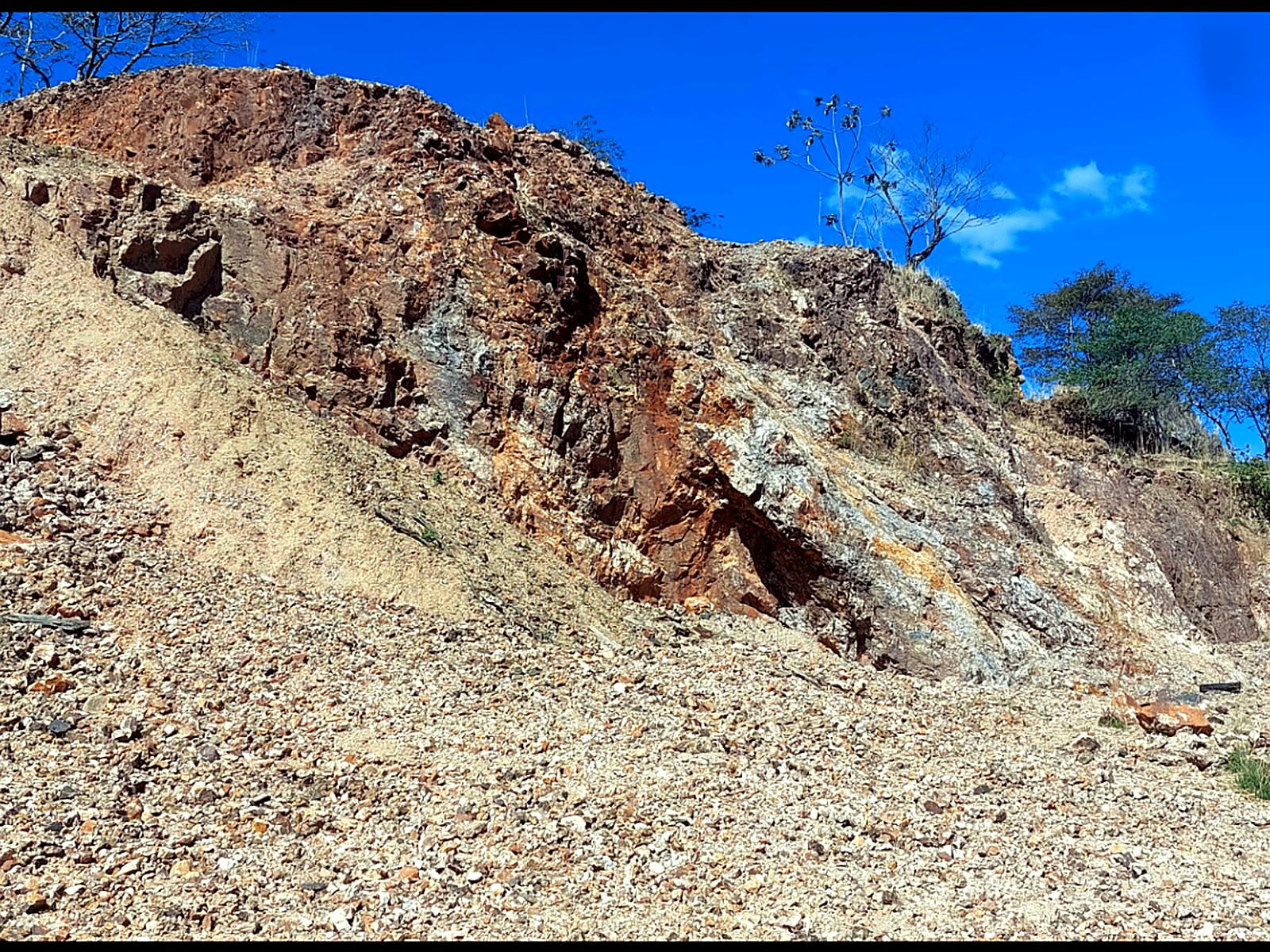 Hillside open cut under blue sky showing a white quartz vein through rust-red oxidised rock