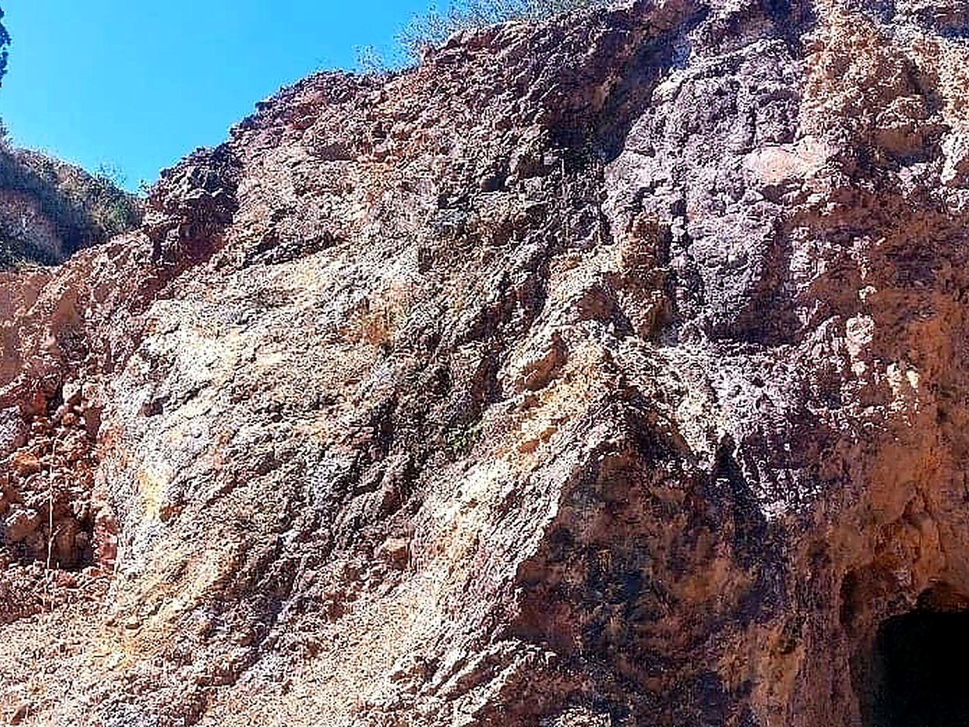 A steep rust-orange and grey oxidised rock face exposing mineralisation against a clear blue sky