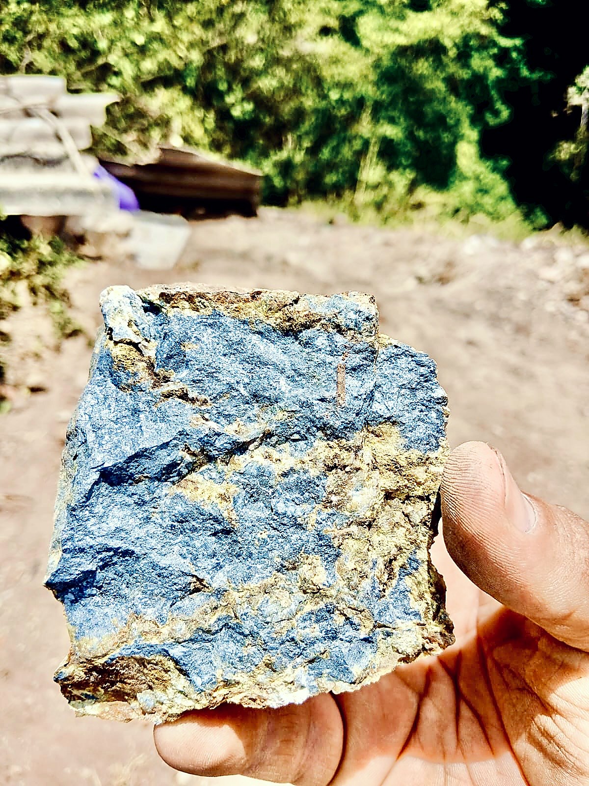 Hand holding a rough rock specimen with a metallic steel-blue antimony face and yellow-brown iron-stained quartz along its edges