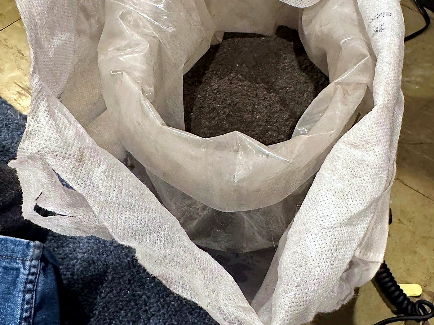 An open white woven sack filled with grey mineral concentrate held open for sampling