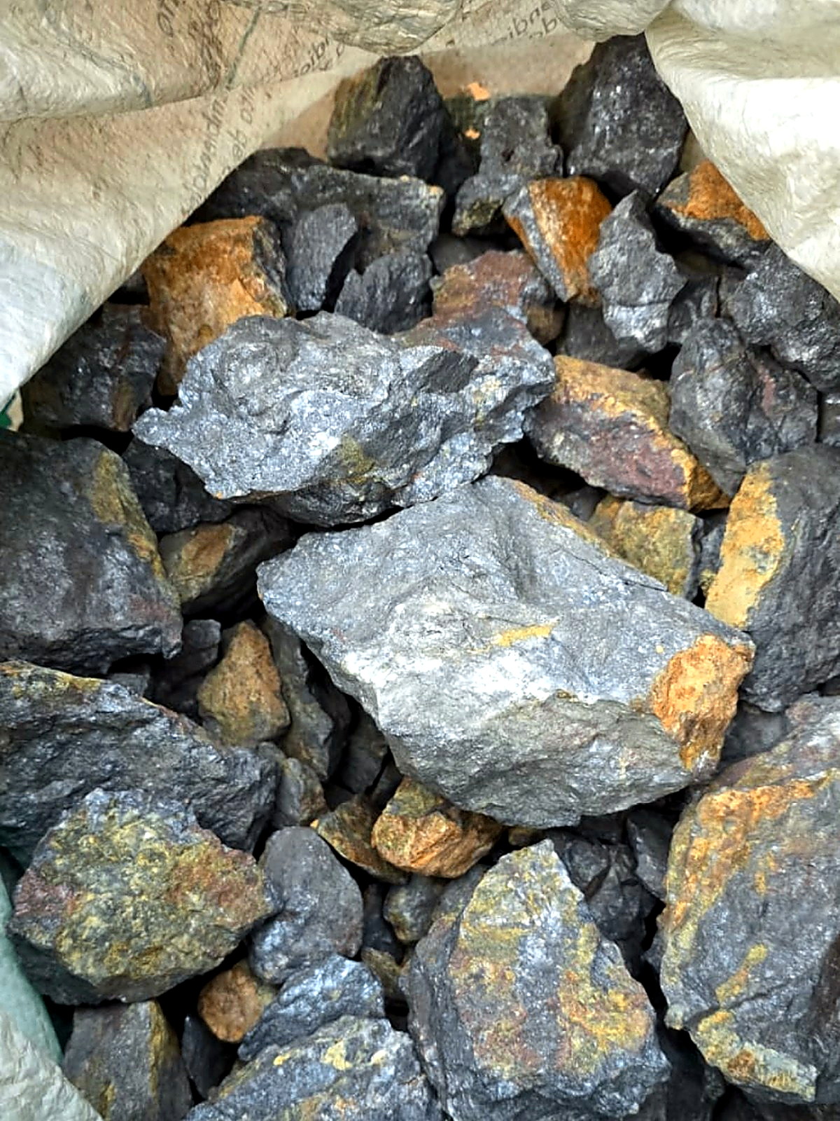 Open woven bulk sack filled with grey crushed sulphide ore chunks streaked with rusty orange oxidation