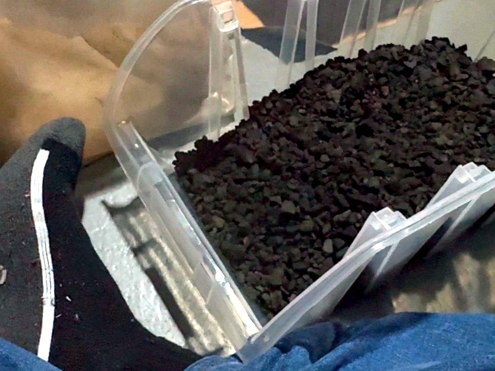 Gloved hands filling a clear plastic tray with dark crushed ore for laboratory sample preparation