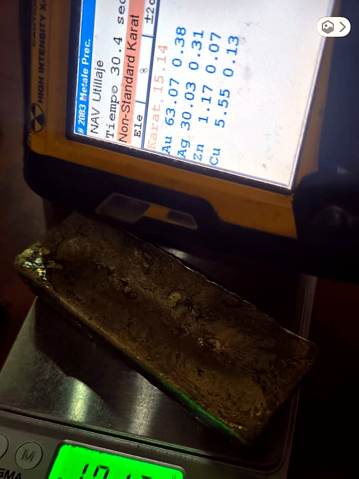 Rough dark gold-silver doré bar in front of a handheld XRF analyser screen listing gold, silver, zinc and copper percentages