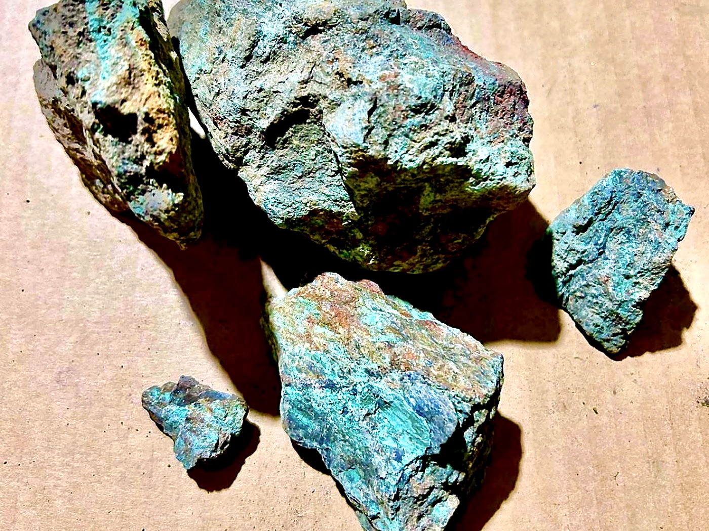 Five pieces of rough copper oxide ore with bright turquoise-green malachite and chrysocolla surfaces on a brown paper sheet