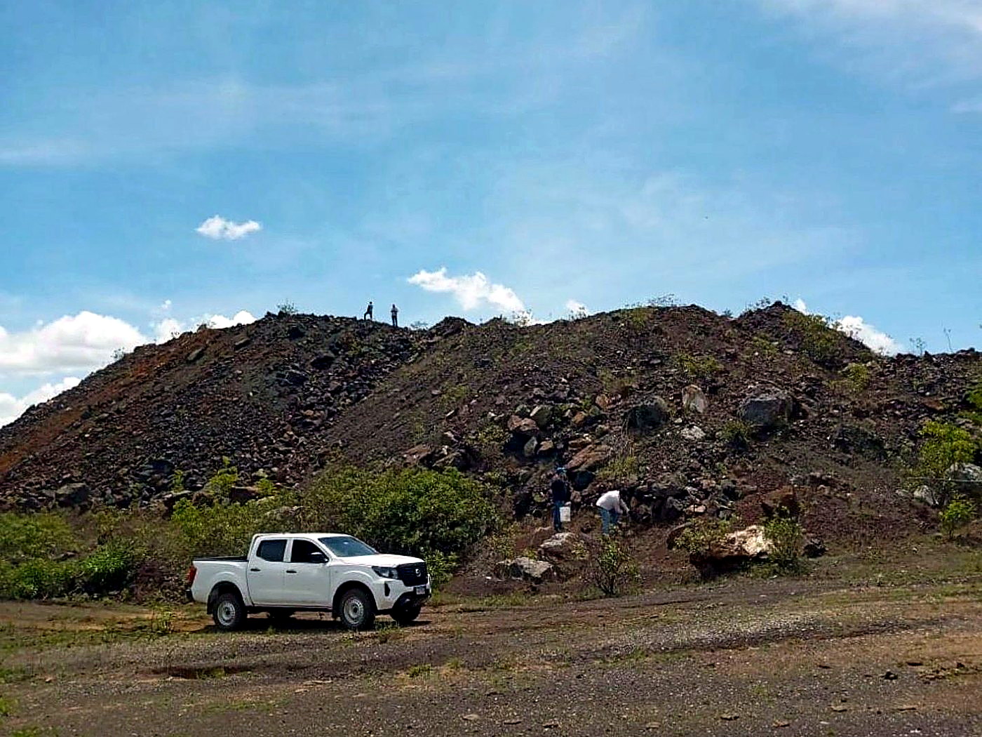 A white pickup truck parked at the base of a dark grey graded ore stockpile on a haulage pad