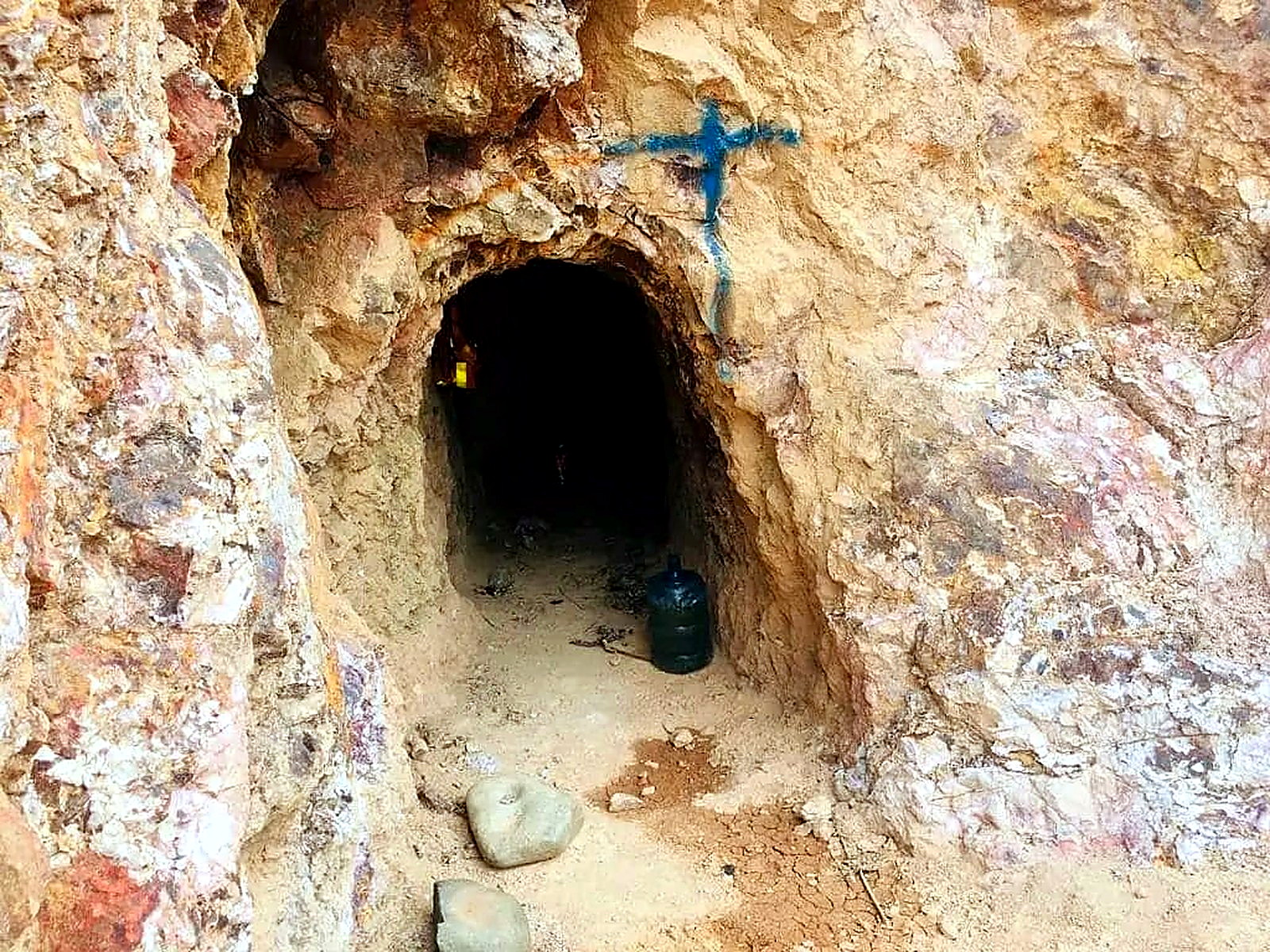 Small arched mine tunnel entrance driven into orange and white oxidised rock, with a blue painted survey cross above the portal