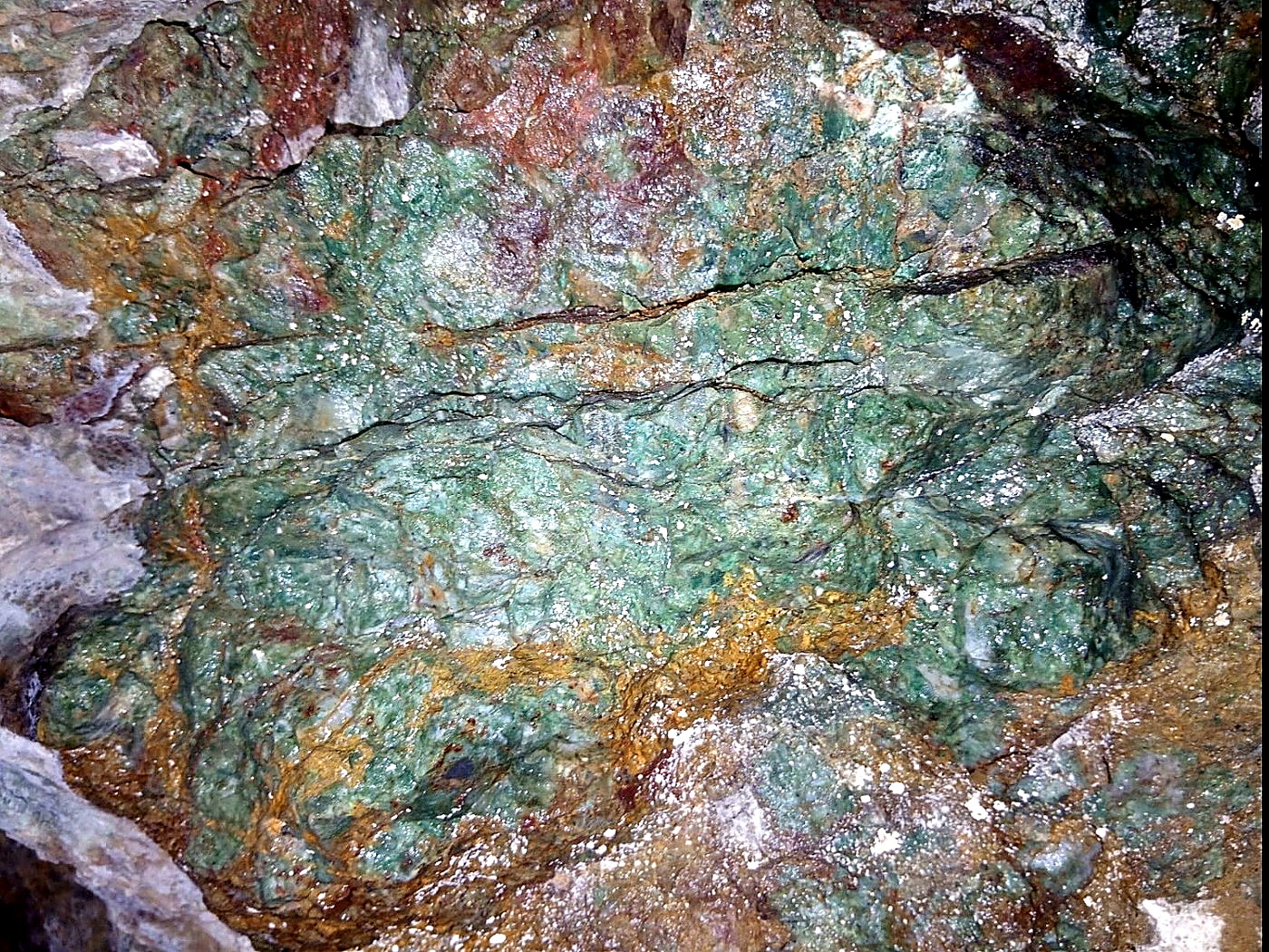 Rock face covered in green malachite and chrysocolla copper mineralisation with orange iron oxide seams and white quartz