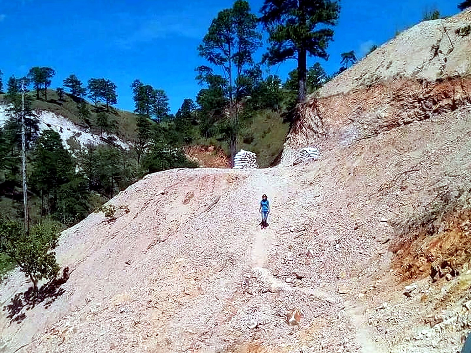 Steep pale rock haul road climbing to a bench where white bulk ore bags are stacked, with a worker walking down